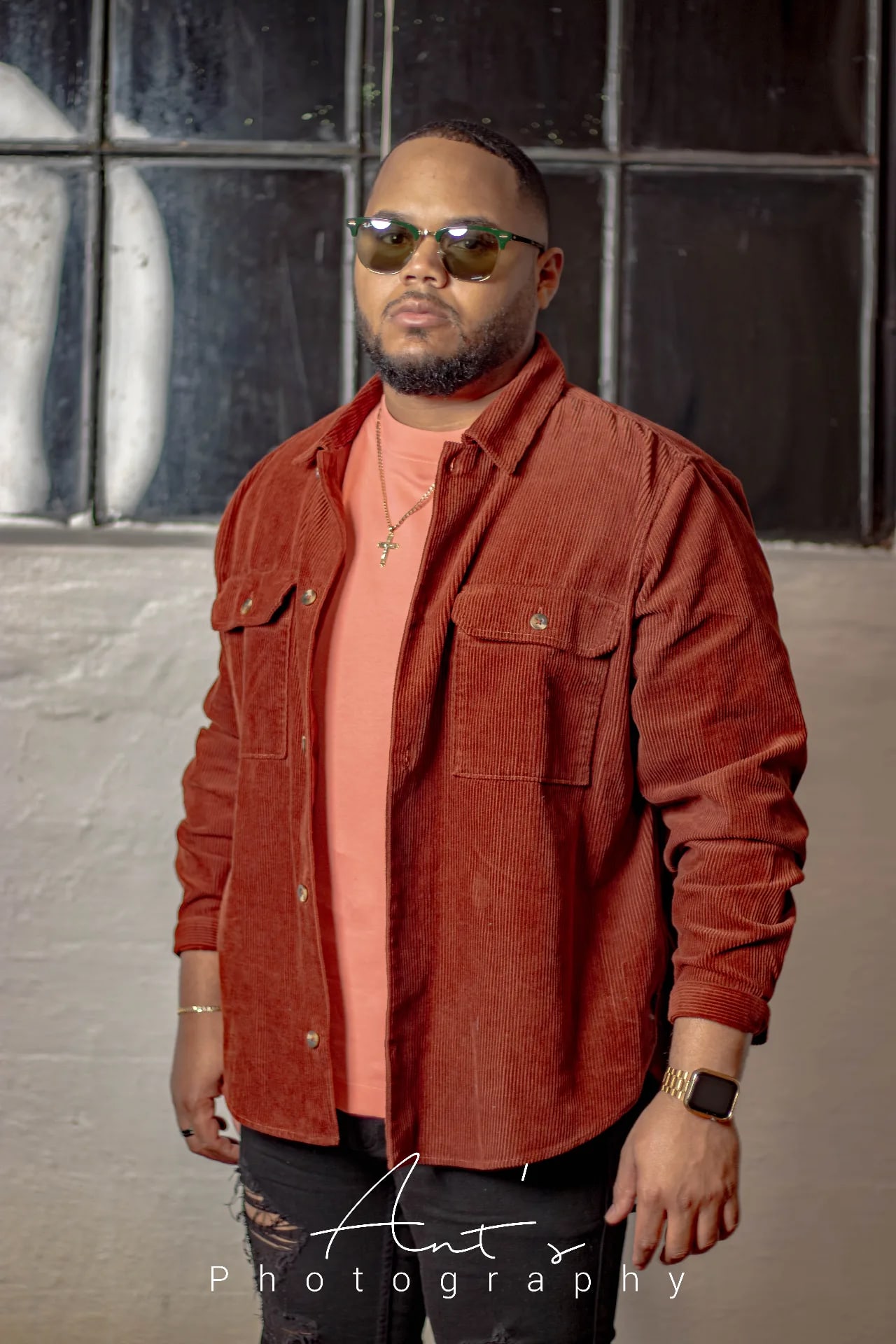 Man in sunglasses and a rust corduroy shirt over a coral tee standing in front of a black-framed window