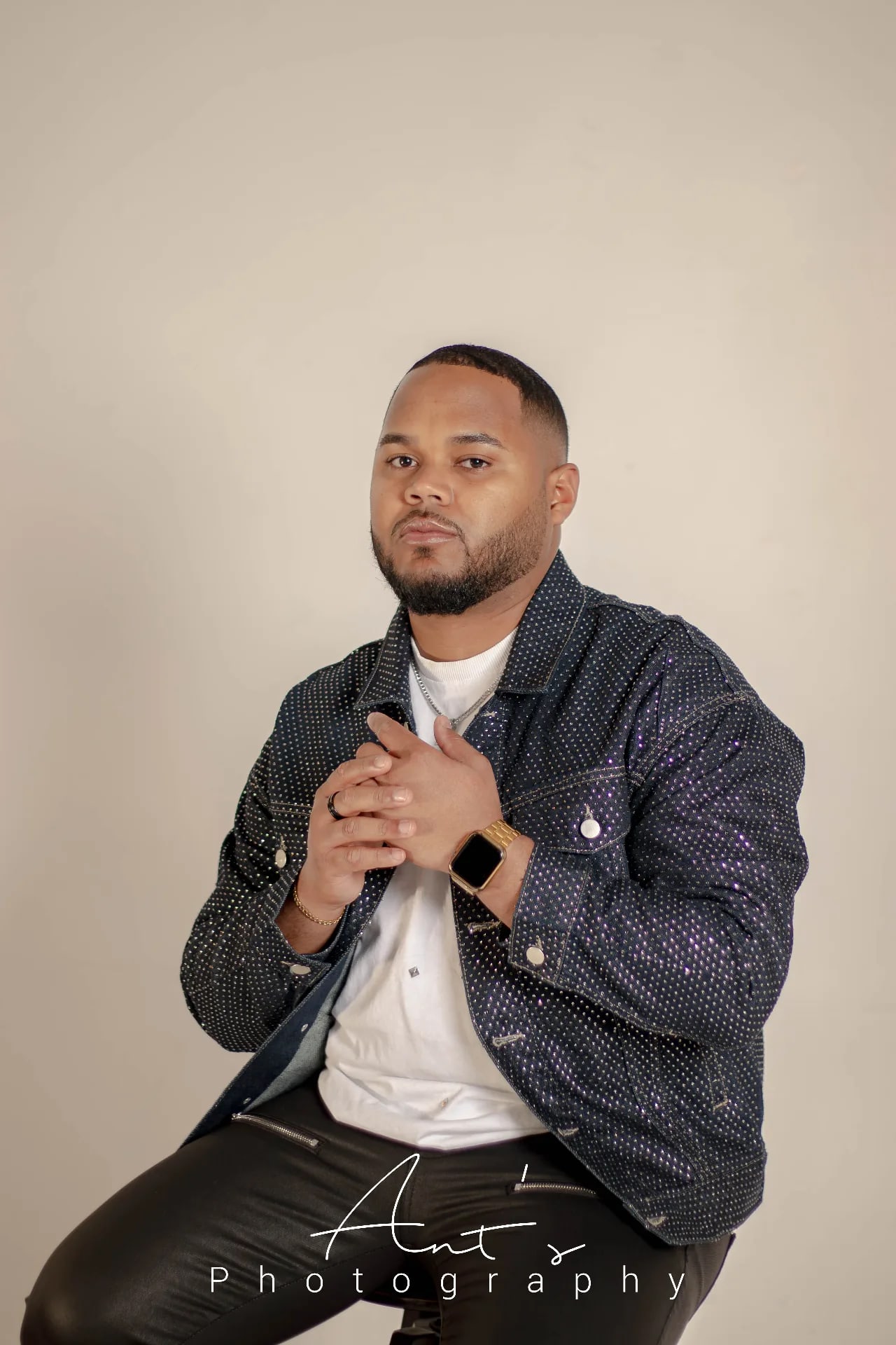 Man in a dark denim jacket seated with his hands clasped in front of a cream studio backdrop